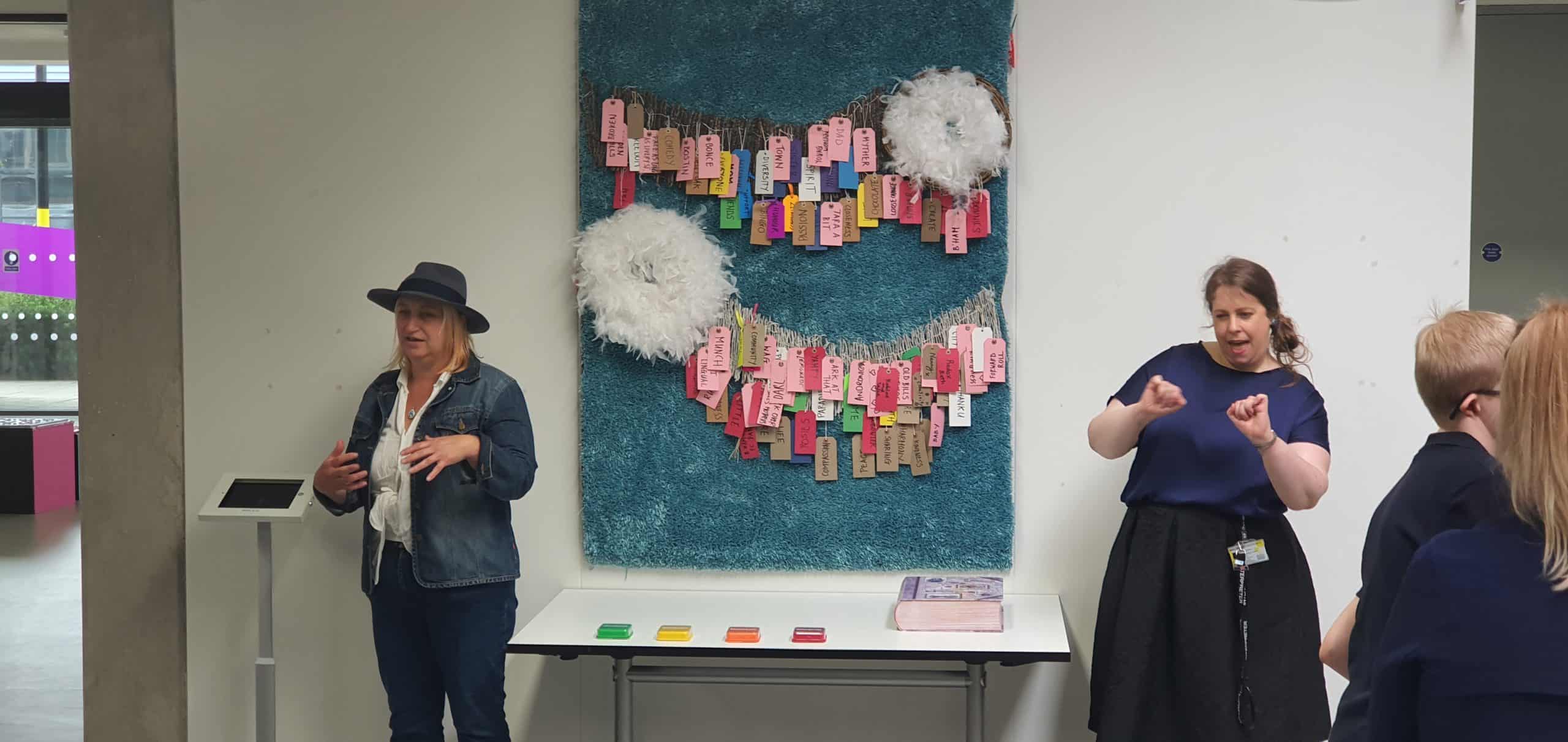 A woman wearing a hat talking about an piece of art hanging on a wall. Art is made of labels