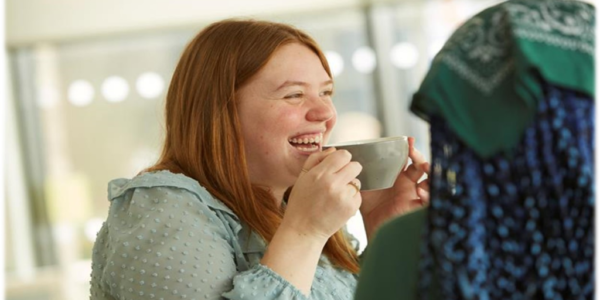 A lady with ginger hair is smiling as she takes a sip of a drink from a teacup.