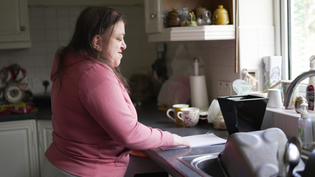 Charlotte, a white woman with brown hair wearing a pink jumper, stands at her kitchen counter looking at a bill.