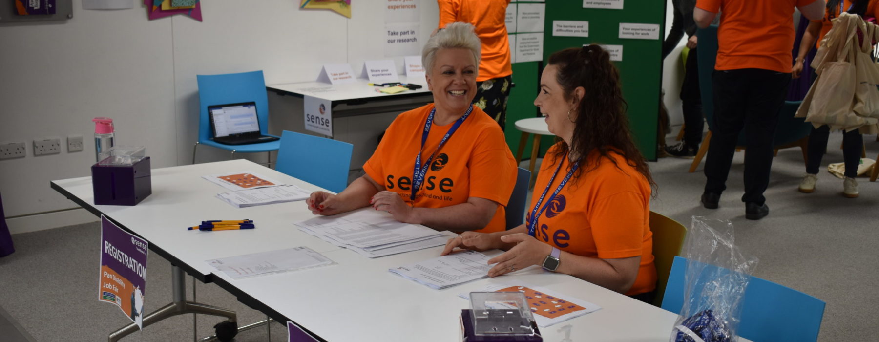 A woman with brown hair and a woman with short blonde hair are sitting at a table wearing bright orange sense t shirts
