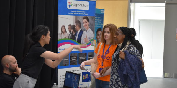 A woman with braids, linking arms with a woman wearing a Sense t-shirt, talks to a woman at a stall at the Pan Disability Jobs Fair.