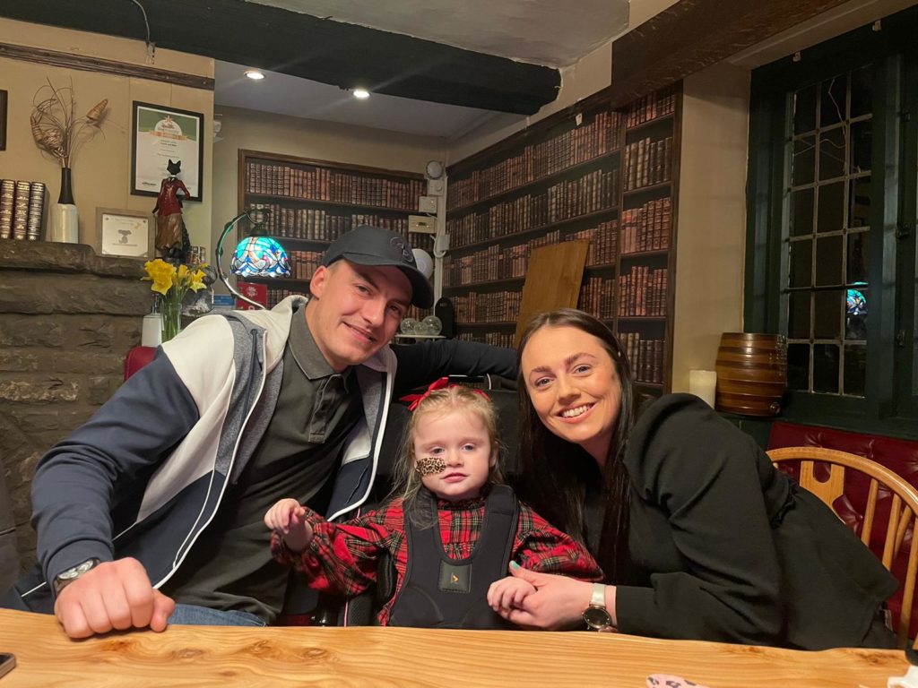 Lowri and Jack and their daughter Mia smile for the camera while sitting at a table together.
