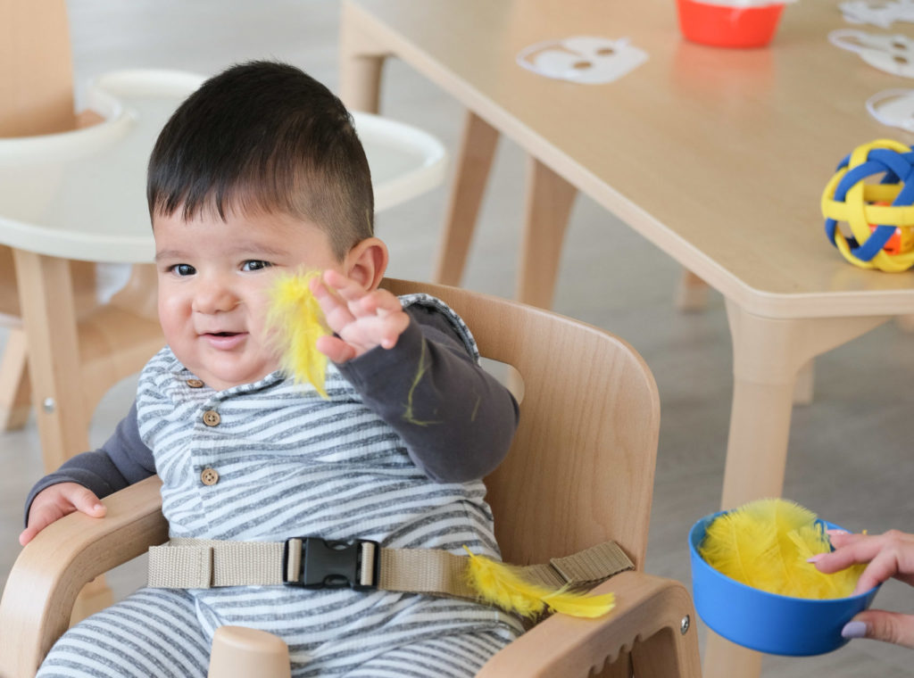 A young boy is sitting in a wooden high chair playing with yellow feathers.