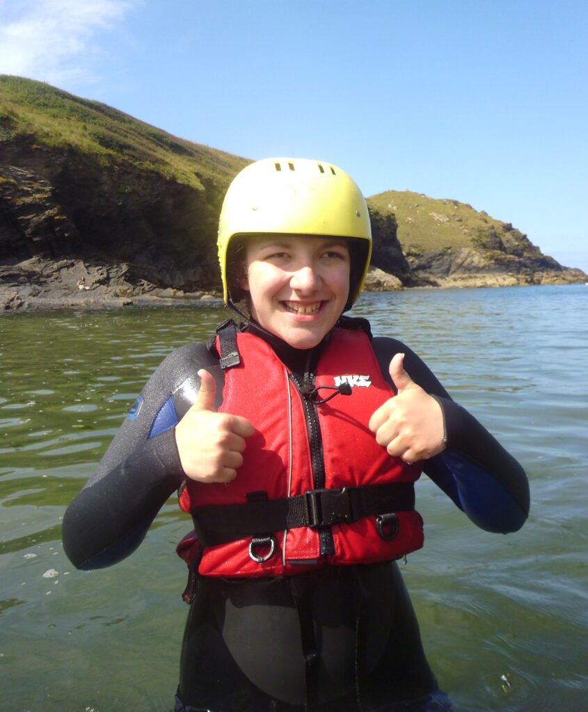 A girl in a lifejacket and helmet smiles and gives a thumbs up