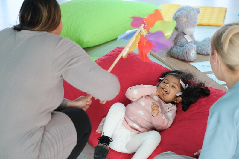 A little girl lying on a cushion enjoys some sensory play, as an adult holds a colourful windmill toy for her to look at.
