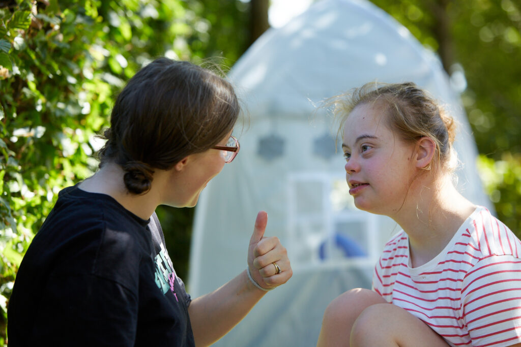 A young girl and volunteer in conversation, with a tent and woodland in the background.