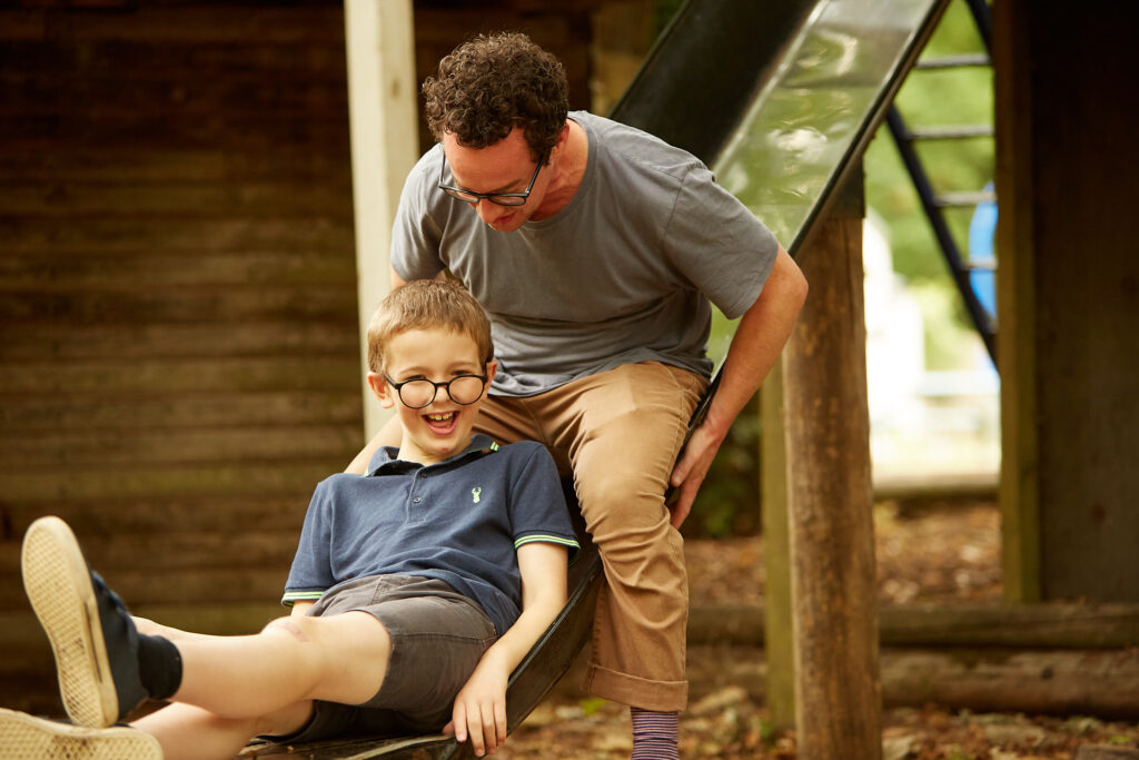 A young boy in glasses laughs while playing on a slide, with an adult male volunteer.