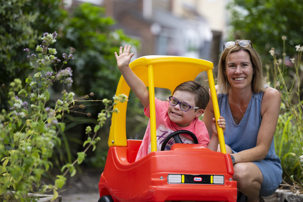 A young boy sitting in a little car in the garden with his mum.