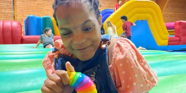 On a big inflatable play area, a girl with braided hair, wearing an orange top and dungarees plays with a colourful toy.