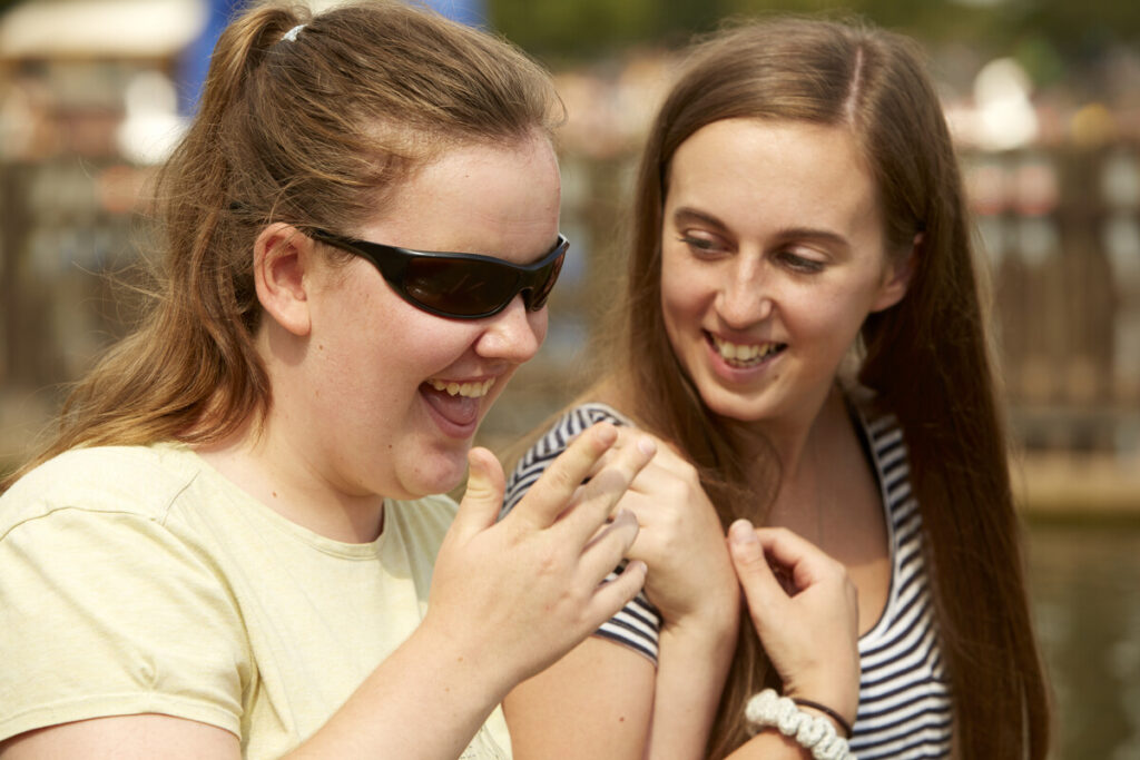 A young woman in sunglasses with her hair in a ponytail, laughing while linking arms with a woman with long brown hair.