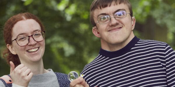 A woman and a man standing and smiling in a park. She is giving a thumbs up and he is holding a flower.