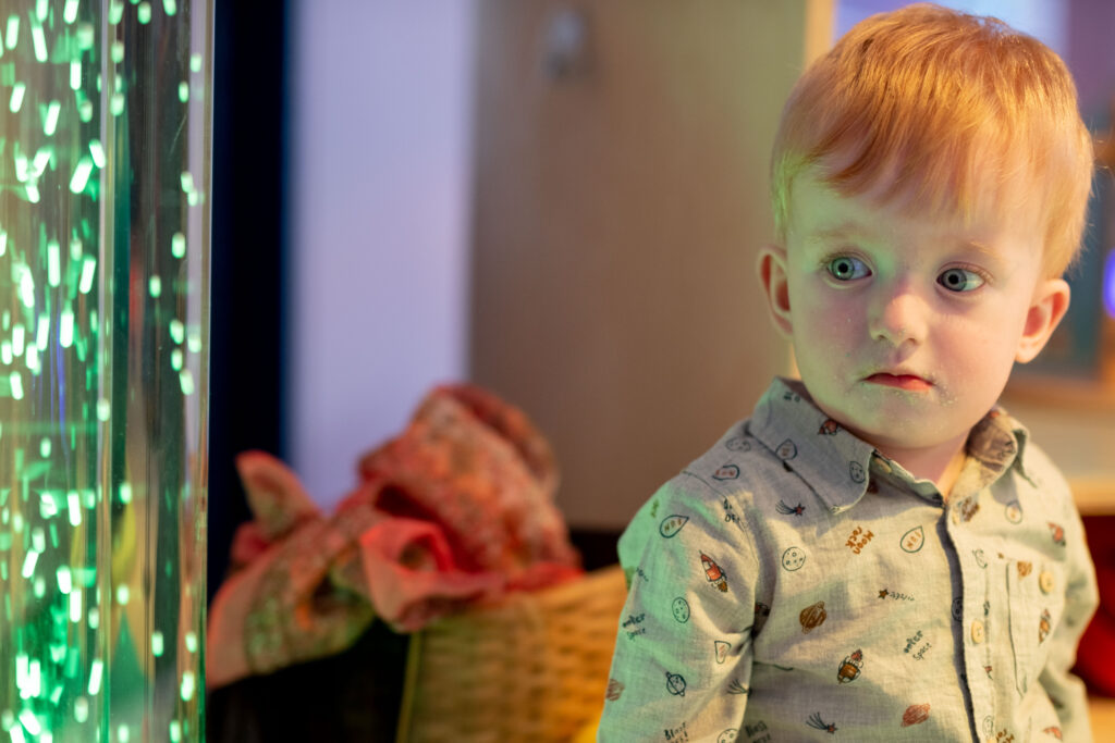 A young boy with blonde hair wearing a shirt is looking at a tube of bubbles illuminated green. 
