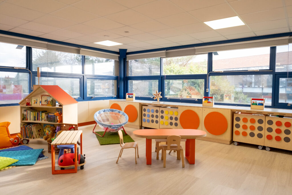 A large spacious room with windows covering all of the walls. There is a row of wooden storage units, covered in giant colourful braille dots along one wall. In front of the storage units is a small orange wooden table with pale wooden chairs, a storage unit shaped like a house filled with booked and colourful mats on the floor. 