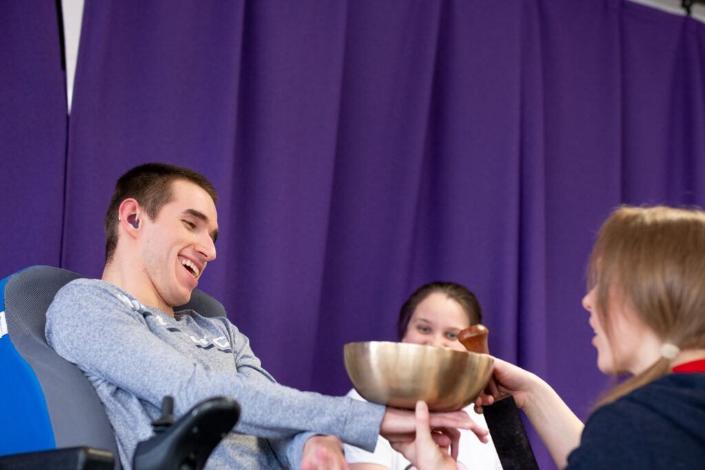 A young man in a chair smiles as he feels the vibrations from a gold singing bowl on his hands. Behind him are dark purple curtains. 