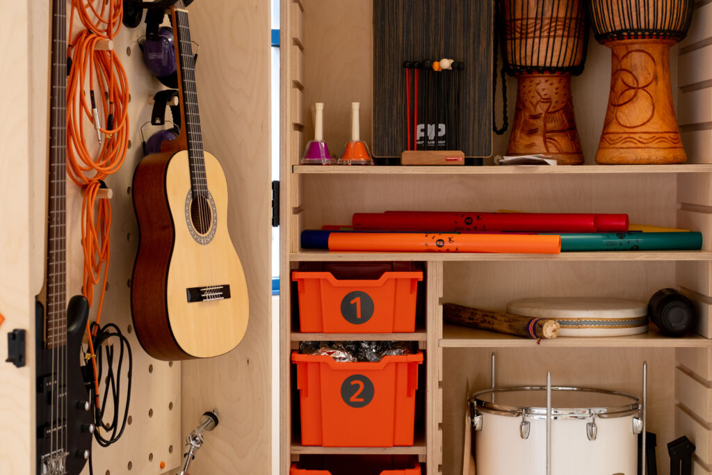 The inside of a wooden cabinet housing lots of music equipment. On the left, the inside of the cabinet door, there are two guitars hanging up, cables wound up and headphones hanging on hooks. On the right are shelves housing bongo drums, colourful bells, drumsticks, colourful tubes and numbered orange boxes. 