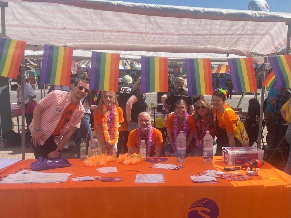 Group of six people in Sense t-shorts smiling behind a large table with leaflets and stickers for Pride