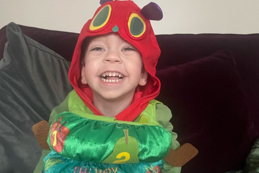 A boy with a massive grin sits on a maroon sofa wearing a very hungry caterpillar costume, including a red hood with yellow eyes on it over his head