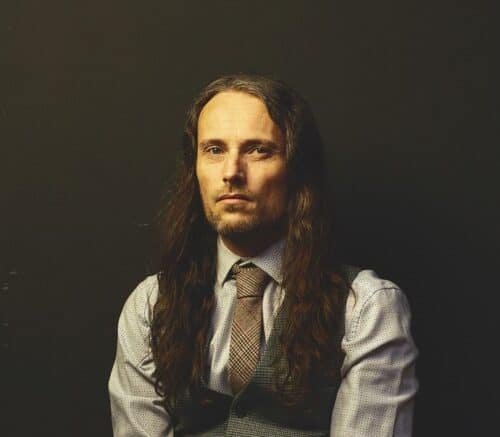 A close-up of Ian Treherne, a white man with long, dark brown hair wearing a shirt, waistcoat and tie.