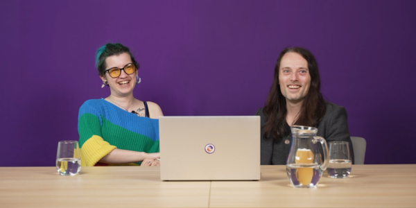 Max, a white genderqueer person with green hair, and Ian, a white man with long dark hair, sitting at a table with a laptop.