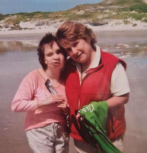 Two women, a mother and daughter are standing on a beach. The daughter has short dark brown hair and is wearing a pink top. The mother has longer, light brown hair and is wearing a cream polo shirt and red gilet.