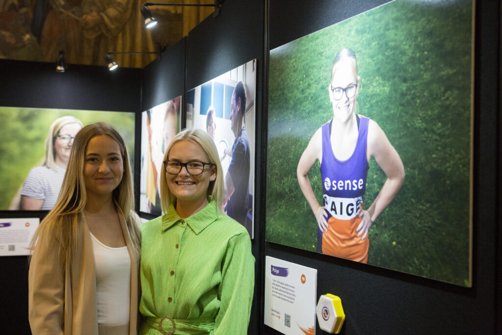 Two blond smiling women looking at the camera.  They are stood next to a photo of one of the women in an exhibition