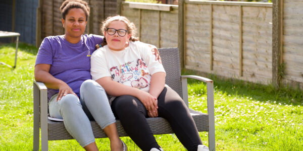 Two young women sit on a bench in a garden, one with her arm around the other.