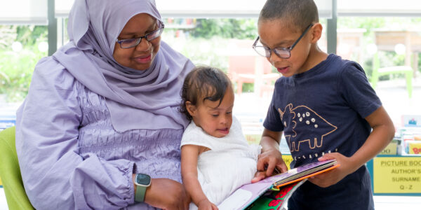 A woman wearing a purple hijab and glasses reads a book with two small children.