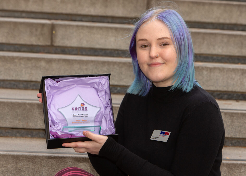 Lauren Gilbert, a person with light blue hair and a nose ring, holding their Sense Award.