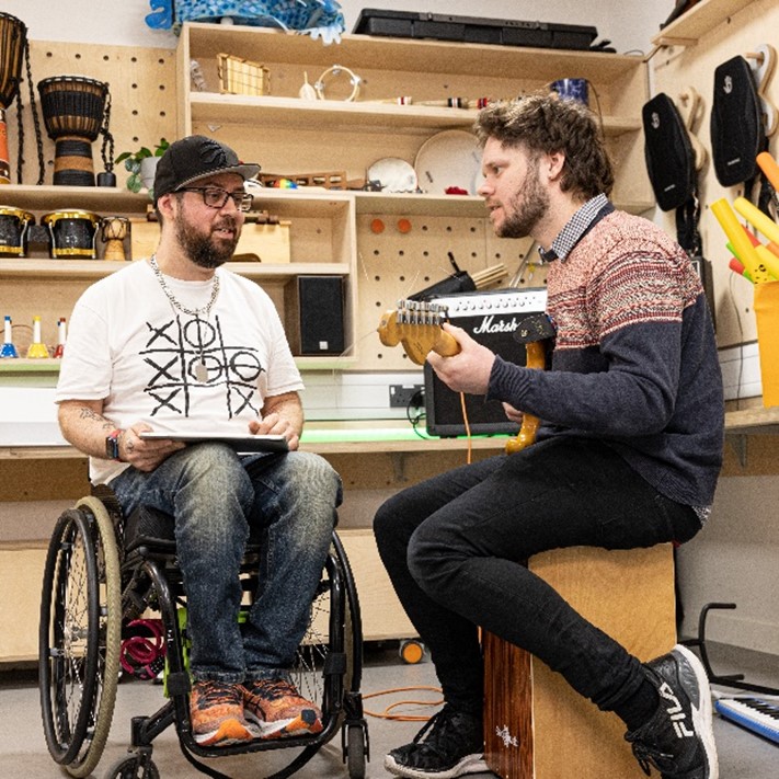 Kris Halpin, a bearded man in a wheelchair, speaking with another man who is playing guitar.