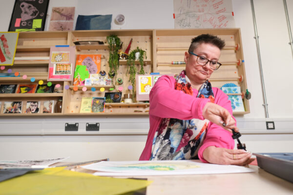 Tanya Raabe Web, Sense's artistic director, sits at a desk working on an artwork in a studio with arts materials in behind her