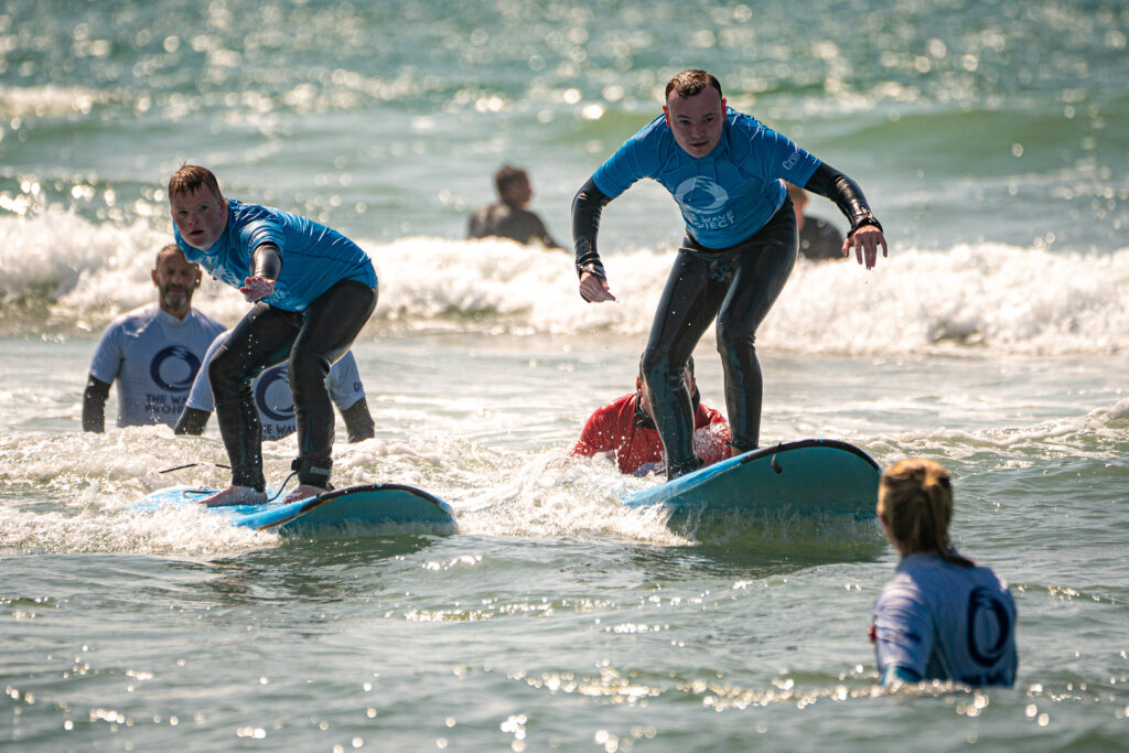 Two young men in wetsuits and blue t-shirts surf along a calm sea filled with people.