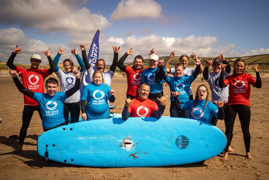 A group of young men and women stand on a beach behind a surf board. They smile at the camera and make the 'hang loose' hand sign.