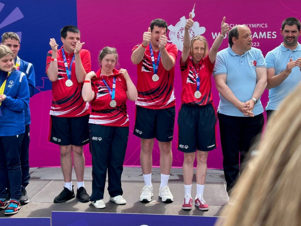 A group of people giving "thumbs up" signs as they stand on stage wearing silver medals.