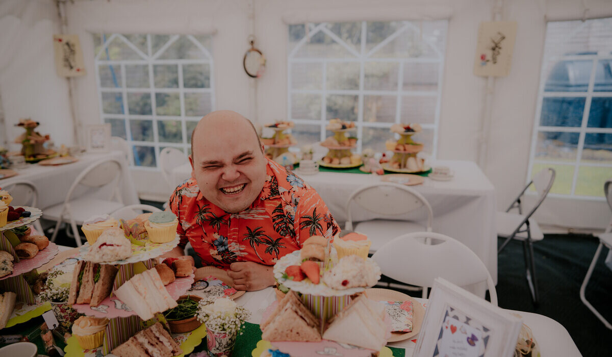 A man sit behind a table piled with cake for afternoon tea, he has a big smile.