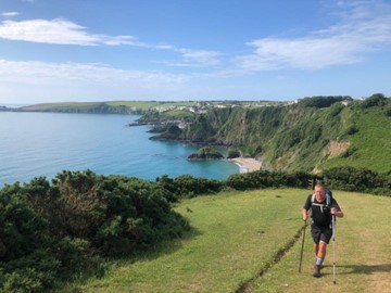 A man walks along a sunny coastal path with the sea and cliffs beside him.