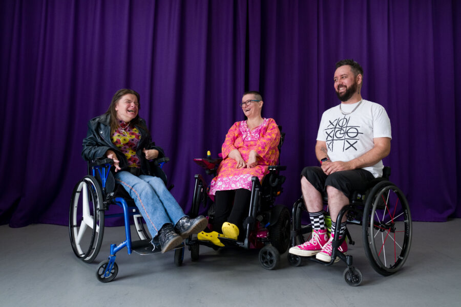 Three people in wheelchairs smiling and laughing in front of a purple curtain