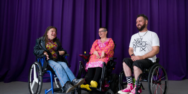 Three people in wheelchairs laughing in front of a purple curtain.