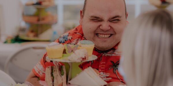 A smiling man sits behind a cake stand piled with sandwiches and cakes.