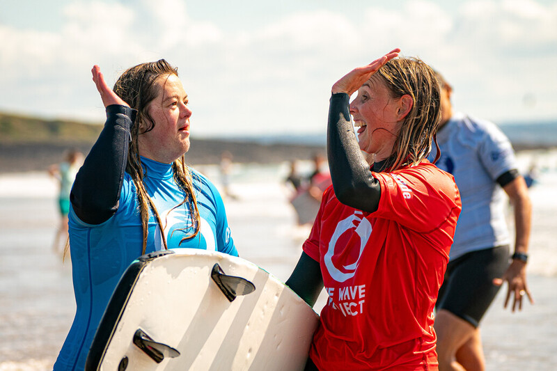 Two women in wetsuits hold surfboards in one hand and high five with the other.