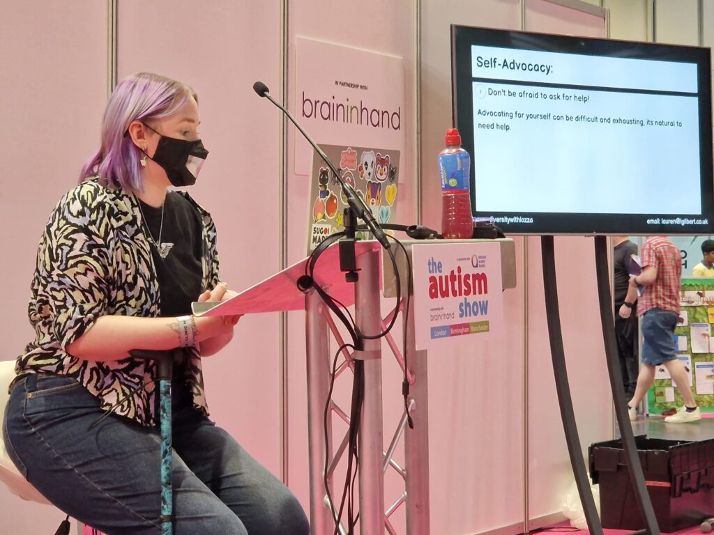 Lauren Gilbert, a white person with purple hair wearing a face mask, giving a talk at The Autism Show.