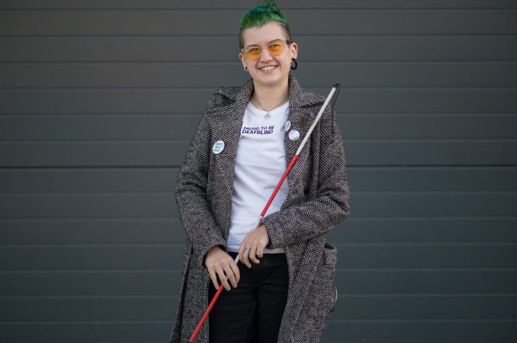 Max, a white genderqueer person with green hair, holding their red and white cane. 