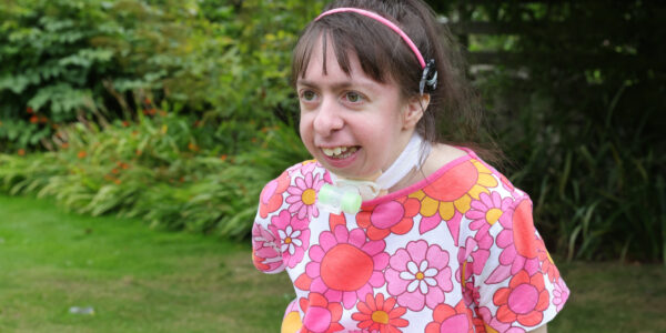 Rebecca, a young white woman with dark hair and a hearing implant on a pink band, smiling in a garden.
