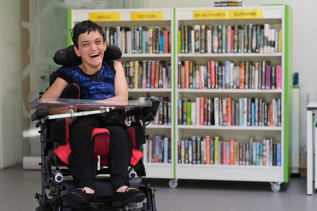 Jade, a white woman with short dark hair in a wheelchair, laughing in front of a bookcase.