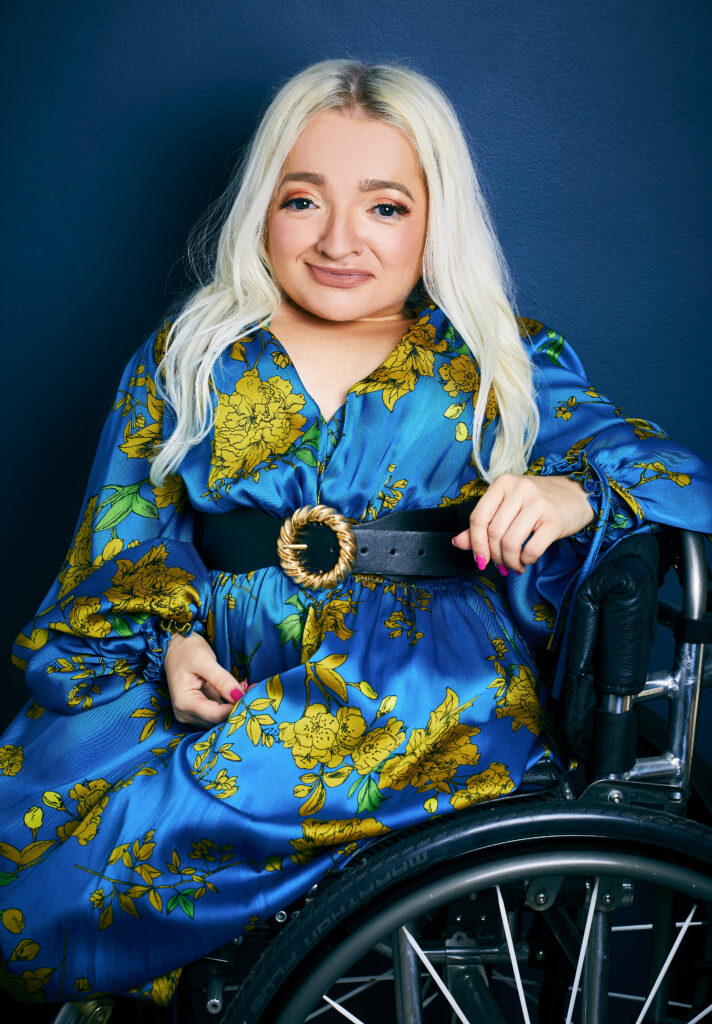 Samantha Renke, a white blonde woman in a blue dress, sitting in her wheelchair.