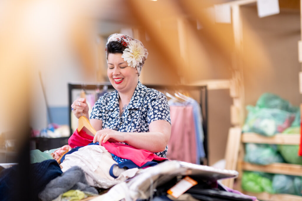 A woman wearing a floral headdress and red lipstick laughs as she sorts through stock at a Sense charity shop.