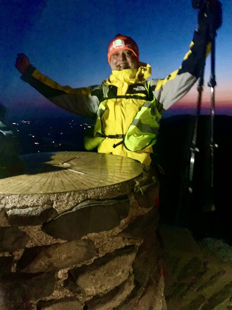 Guni Suri at the peak of Snowdon in the middle of the night, lit by torchlight in the darkness, arms raised in celebration.