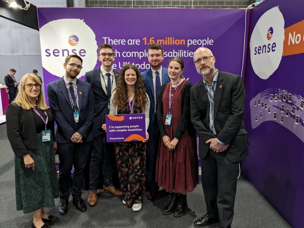 Image of 7 people stood in a line in front of a Sense branded background. Sarah (a woman with brown curly hair) is in the middle holding a sign that says 'I'm supporting people with complex disabilities'