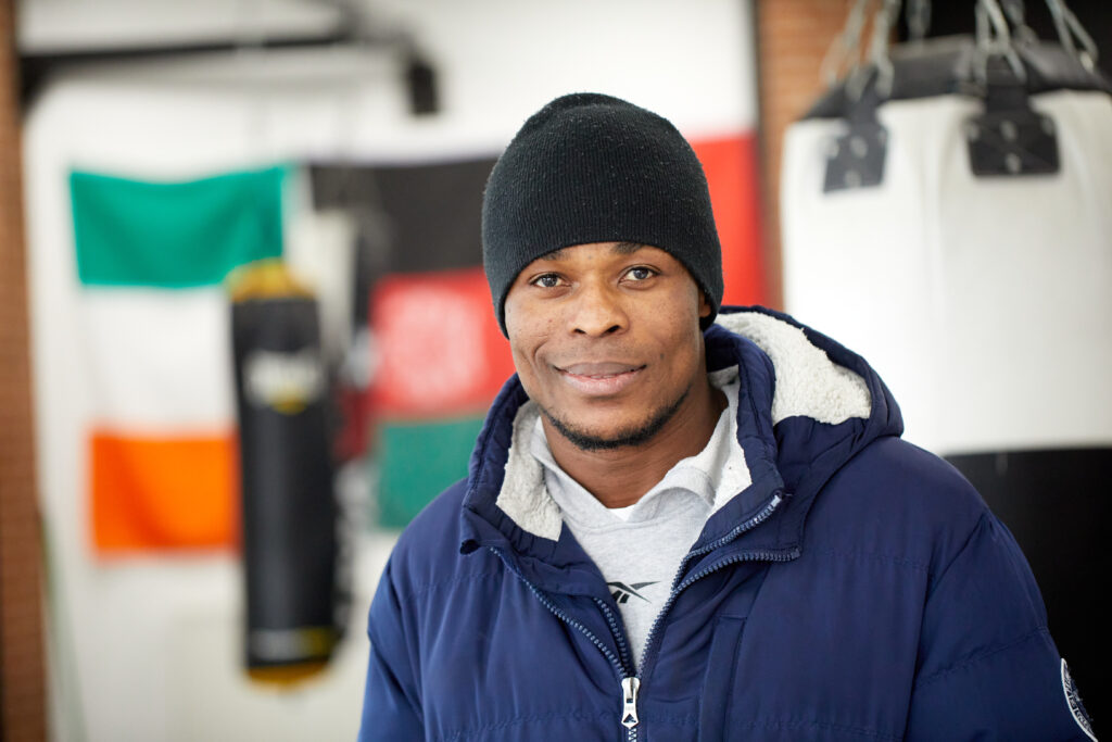 A young smiling man wearing a coat and beanie stands inside a boxing centre.