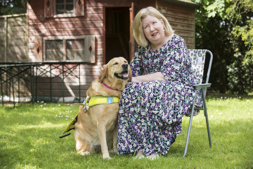 Jane Manley, a blonde woman wearing a floral dress, sitting in her garden with her guide dog.
