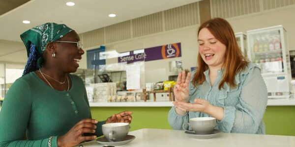 Two ladies sign language with a mug and saucer in front of them in a cafe.